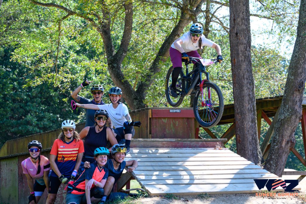 Mountain biker riding off a drop with a group of ladies standing nearby.
