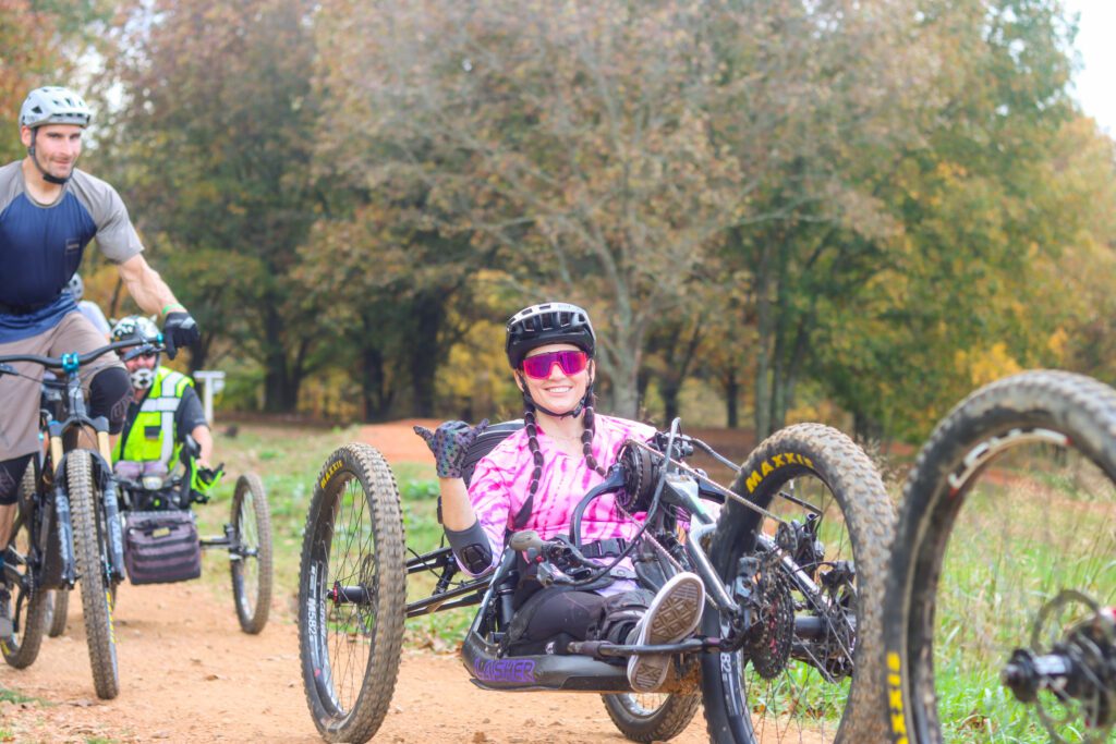 A female adaptive mountain biker smiles for a photo as she rides to the top of a downhill trail.